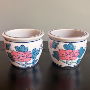 May include: Two small white ceramic planters with a rounded shape and a blue rim. Each planter features a hand-painted floral design with large pink peony blossoms and green leaves wrapping around the exterior of the white porcelain body.