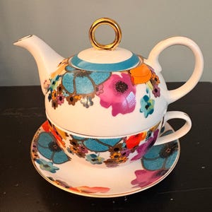 May include: A white ceramic tea set with a floral design. The set includes a teapot with a gold handle, a teacup, and a saucer. The floral pattern features blue, pink, orange, and purple flowers. The set is designed for one person.