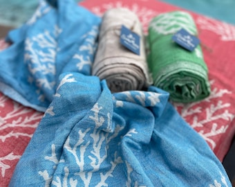 May include: Three rolled up beach towels with a coral reef design. The towels are in shades of blue, green, and beige. The towels are laid out on a red and white patterned beach towel.