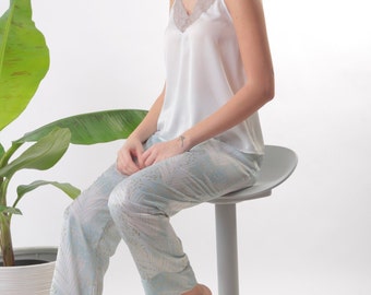 May include: A woman wearing a white satin top with lace trim and light blue patterned pants sits on a gray stool.