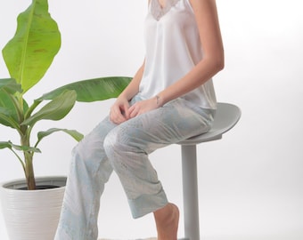 May include: A person wearing a white satin top with lace trim and light blue patterned pants sits on a gray stool. The person's feet are bare and the stool has a round seat.