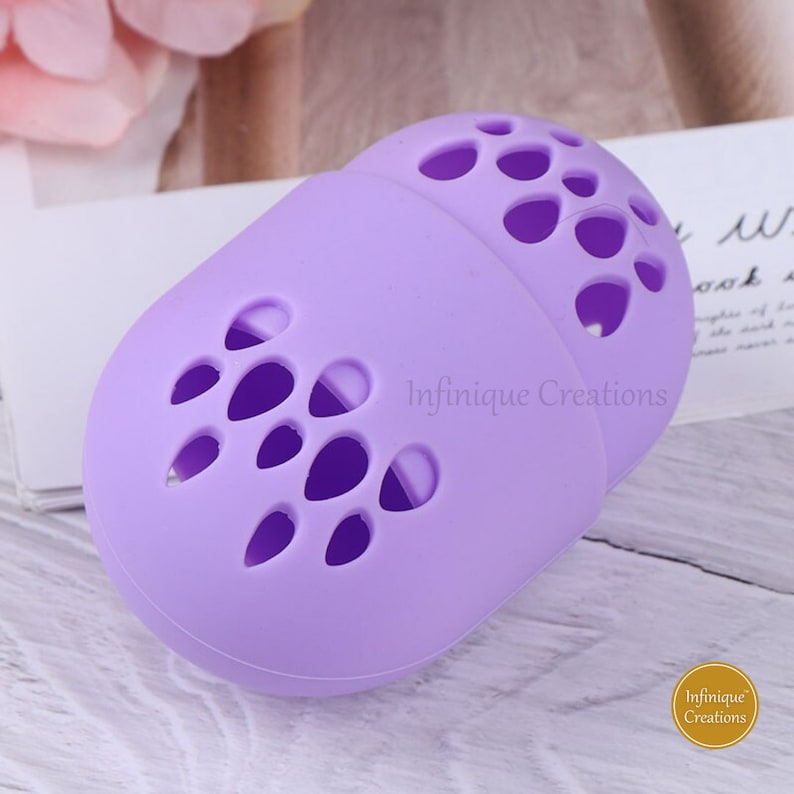 May include: Purple silicone makeup sponge holder with a  pattern of small holes. The holder is shaped like an egg and has the text "Infinifique Creations" printed on the side.