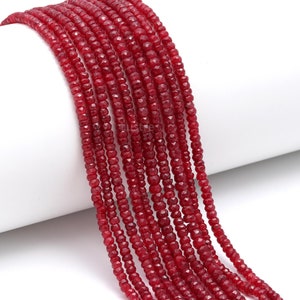 Puede incluir: Una hebra de cuentas de piedras preciosas rojas, facetadas y redondas, enhebradas en una superficie blanca. Las cuentas son pequeñas y de tamaño uniforme.