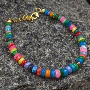 Puede incluir: Una pulsera de cuentas coloridas con un cierre dorado. La pulsera presenta pequeñas cuentas facetadas en tonos de rojo, rosa, amarillo, verde, azul y morado. La pulsera se muestra sobre una superficie texturizada de color gris oscuro.