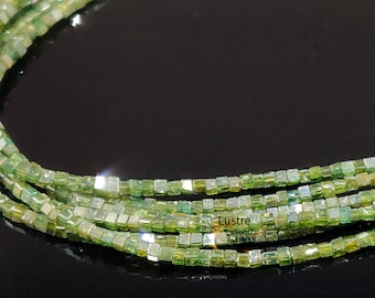 May include: A strand of green gemstone beads, each bead is a square shape with a shiny, reflective surface. The beads are arranged in a line, with the ends of the strand visible. The beads are on a black surface.