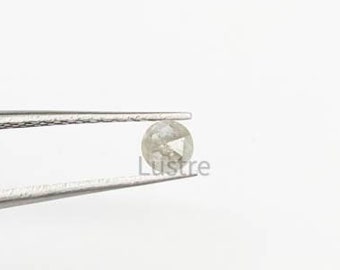 May include: A small, round, light-colored gemstone is held by a pair of silver tweezers against a white background. The gemstone has a faceted surface. The word "Lustre" is printed on the right side of the gemstone.