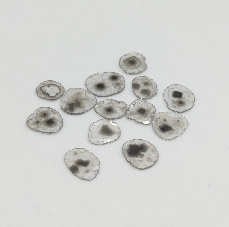 May include: A collection of small, clear, flat, round stones with black inclusions. The stones are scattered on a white surface.
