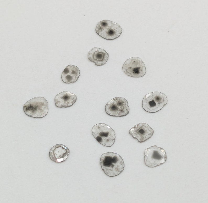 May include: A collection of 14 small, round, clear glass pieces with black and gray inclusions. The pieces are scattered on a white surface.