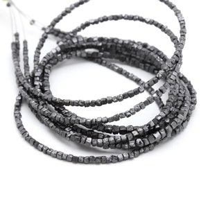 Pode incluir: Um fio de contas de diamante preto, cada conta é um pequeno cubo áspero. As contas são enfiadas em um fio e estão dispostas em uma espiral solta.