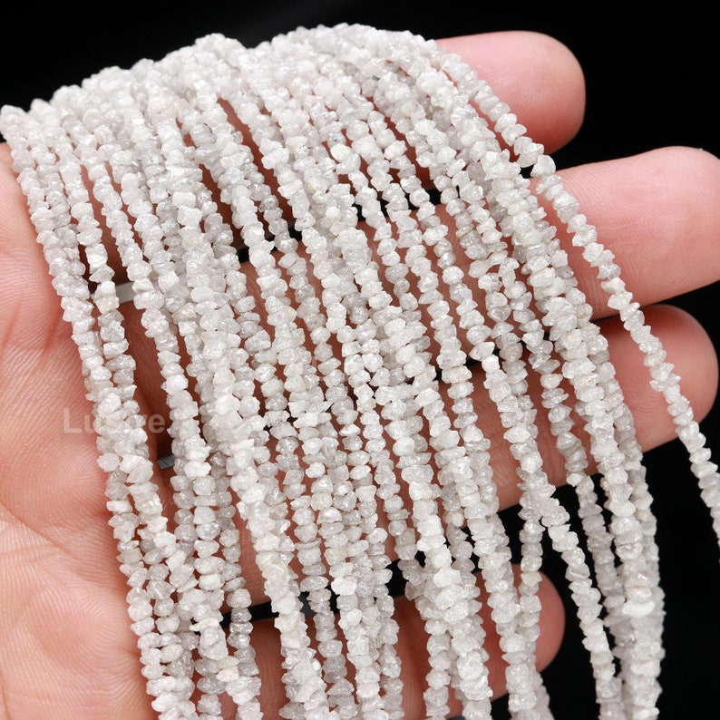 May include: A strand of white rough diamond beads. The beads are small and irregular in shape, and they are strung together on a clear thread. The beads are arranged in a loose, flowing pattern.