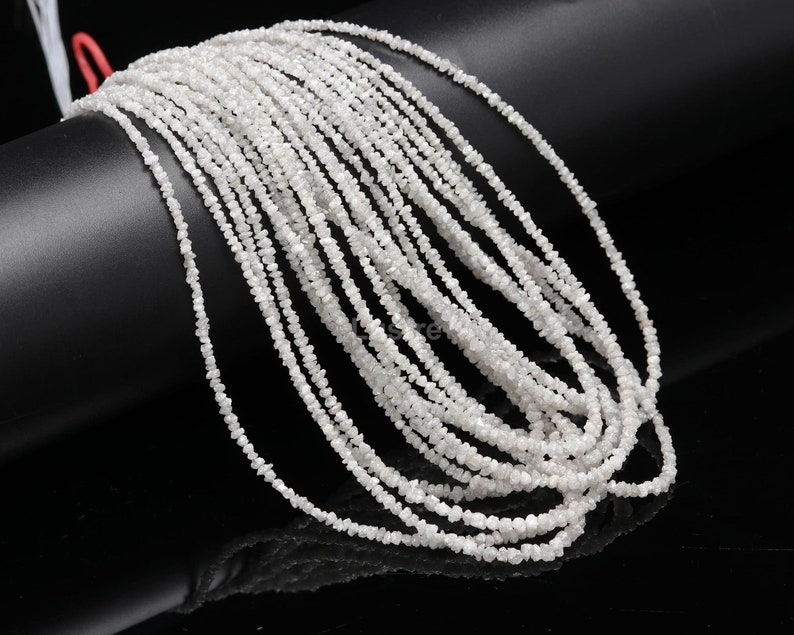May include: A strand of white rough diamond beads. The beads are small and irregular in shape, and they are strung together on a clear thread. The beads are arranged in a loose, flowing pattern.