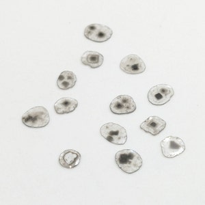 May include: A collection of 13 small, round, clear glass stones with black and gray markings. The stones are scattered on a white surface.