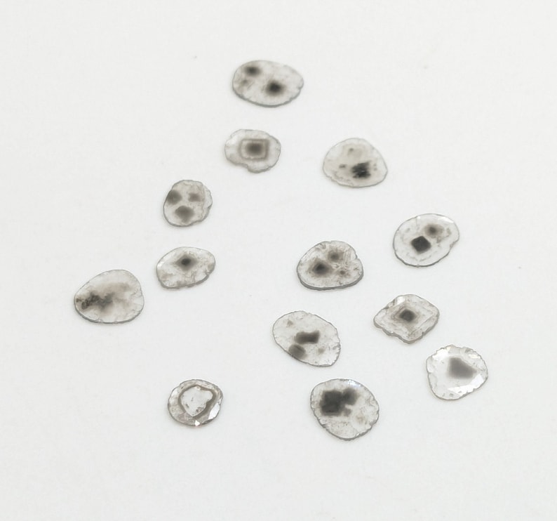 May include: A collection of 13 small, round, clear glass stones with black and gray markings. The stones are scattered on a white surface.