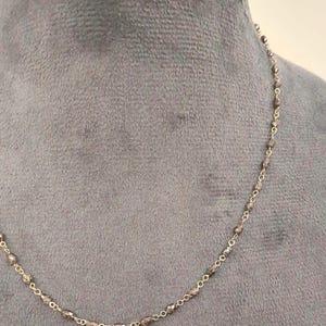 Può includere: Delicata collana color oro con piccole perline grigie sfaccettate. Le perline sono uniformemente distanziate lungo una sottile catena, creando un design minimalista ed elegante. La collana è esposta su uno sfondo grigio, evidenziando la sua sottile lucentezza e la sua estetica raffinata.