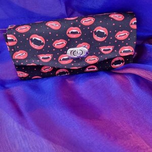 May include: A black clutch bag with a repeating pattern of red lips with fangs on a black background. The bag has a silver clasp.