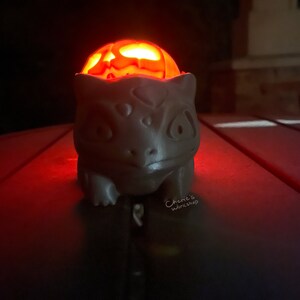 May include: A gray, ceramic Bulbasaur figure with a carved pumpkin head glowing orange inside. The figure is on a dark surface, illuminated by the pumpkin's light. The text "Cherie's Workshop" is visible.