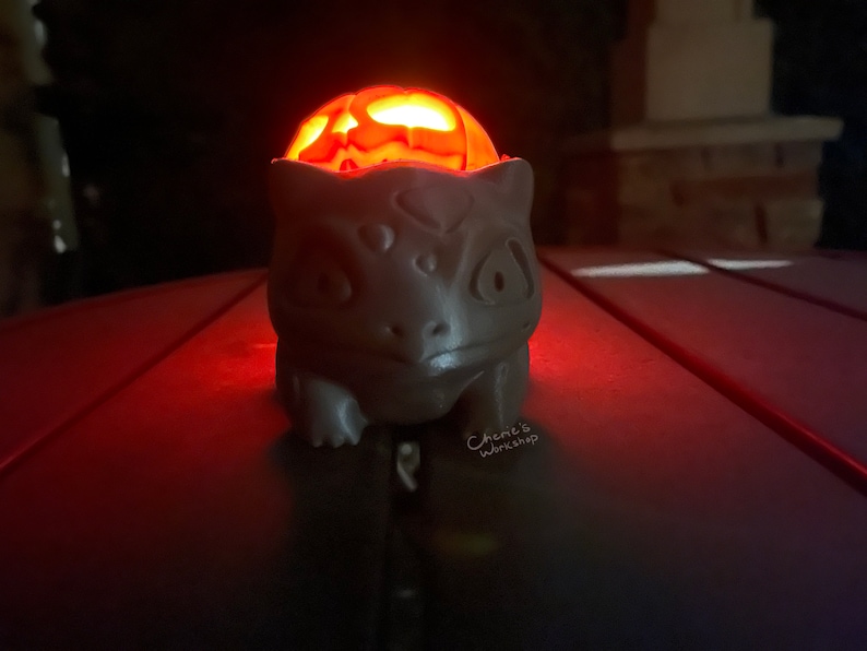May include: A gray, ceramic Bulbasaur figure with a carved pumpkin head glowing orange inside. The figure is on a dark surface, illuminated by the pumpkin's light. The text "Cherie's Workshop" is visible.