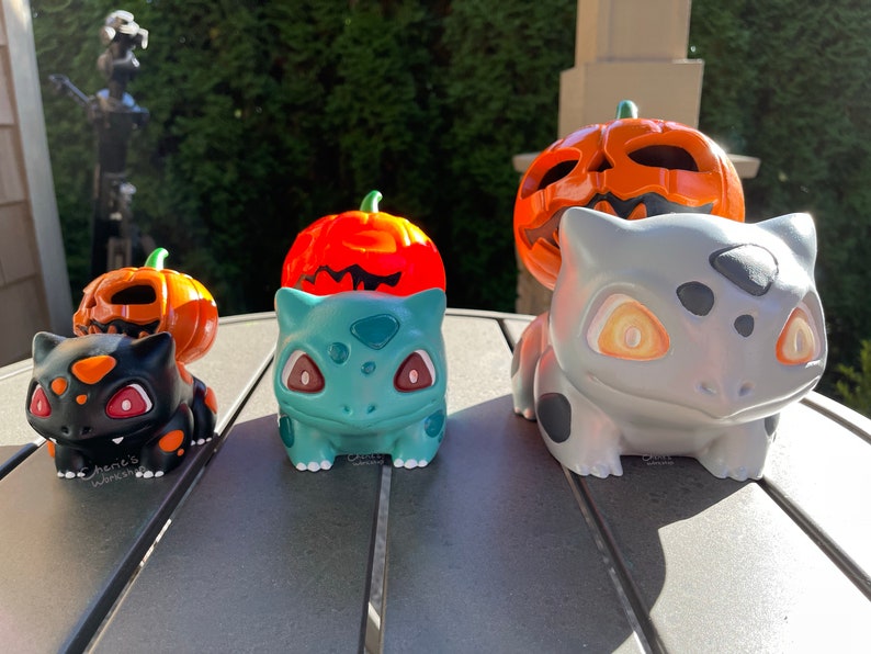 May include: A collection of Bulbasaur figurines with pumpkin-themed tops. The figurines are in black, teal, and gray, with orange and red accents. The figurines are on a dark gray table, with a blurred green background.