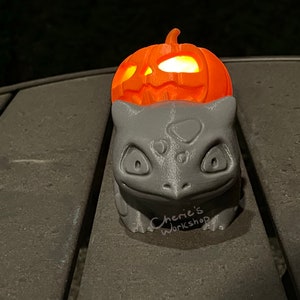 May include: A gray Bulbasaur figure with a carved orange pumpkin on top, illuminated from within. The pumpkin has carved eyes and a mouth. The base of the figure has the text "Cherie's Workshop".