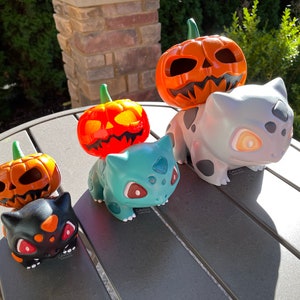 May include: A collection of Halloween-themed figurines. The figurines include carved orange pumpkins and Bulbasaur characters in various colors, including black, teal, and gray. The pumpkins have green stems and carved faces.