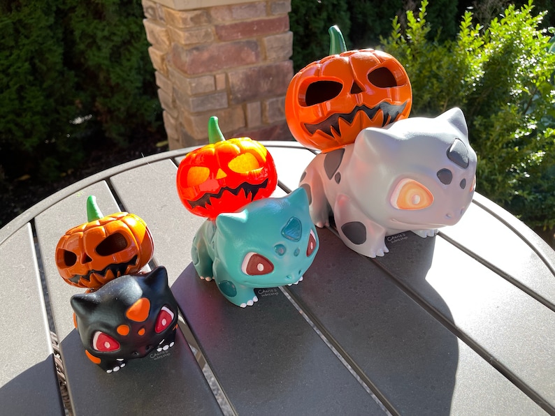 May include: A collection of Halloween-themed figurines. The figurines include carved orange pumpkins and Bulbasaur characters in various colors, including black, teal, and gray. The pumpkins have green stems and carved faces.