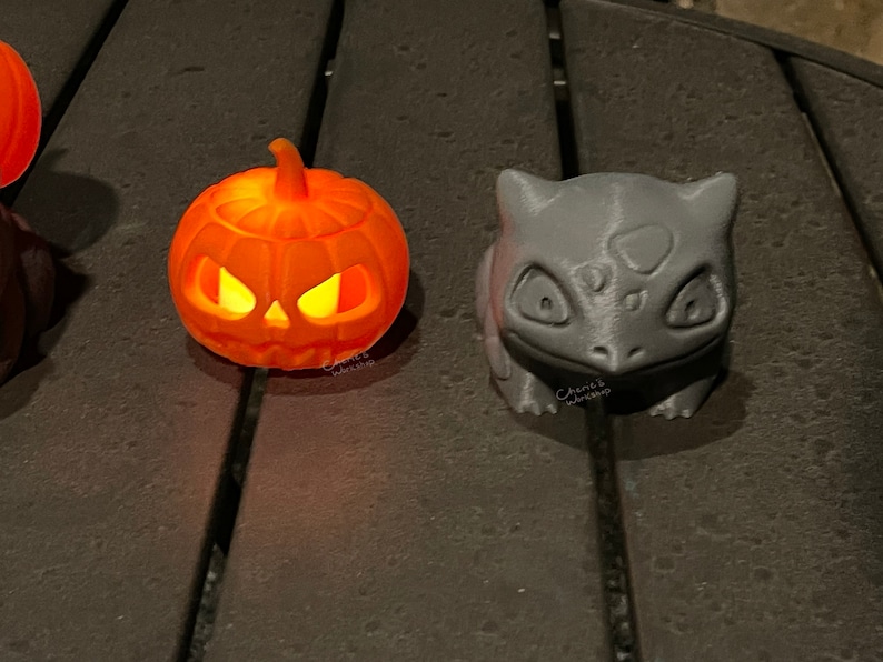 May include: Orange 3D-printed jack-o'-lantern with glowing eyes and a gray 3D-printed Bulbasaur figure. The jack-o'-lantern is lit from within. The Bulbasaur has a detailed, textured surface. Both are Halloween-themed decorations.