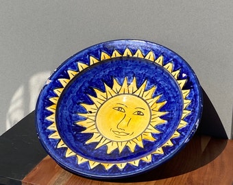 May include: A blue and yellow ceramic plate featuring a sun face design. The sun has a yellow face with closed eyes and a smile. The plate has a blue background with a yellow border.
