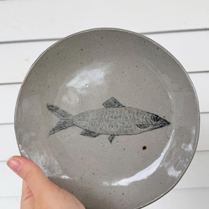 May include: A round, speckled gray ceramic plate featuring a hand-drawn illustration of a fish. The fish is depicted in a detailed, grayscale style, with visible scales and fins. The plate has a slightly raised edge.