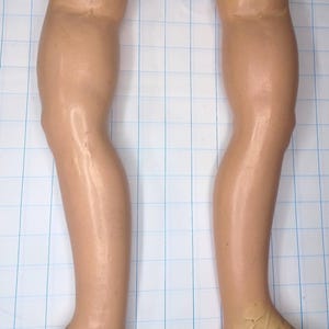 May include: Two vintage doll legs made of a flesh-toned material. The legs are slightly bent at the knees and have a worn, aged appearance. The feet are a lighter color, and the tops of the legs have open cavities.