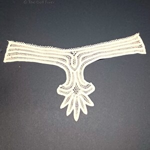 May include: A white lace trim with a scalloped edge and a delicate floral design. The trim is shaped like a Y and has a pointed bottom.