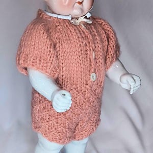 May include: A vintage porcelain doll with painted facial features, blue eyes, and dark hair, wearing a hand-knitted, short-sleeved, peach-colored romper with two white buttons down the front and a small white ribbon collar at the neck.