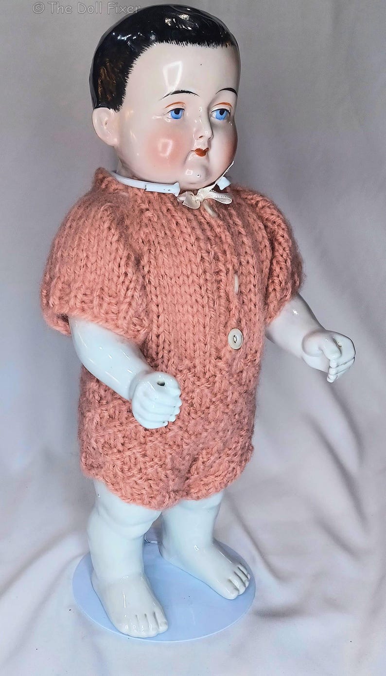 May include: A vintage porcelain doll with painted facial features, blue eyes, and dark hair, wearing a hand-knitted, short-sleeved, peach-colored romper with two white buttons down the front and a small white ribbon collar at the neck.