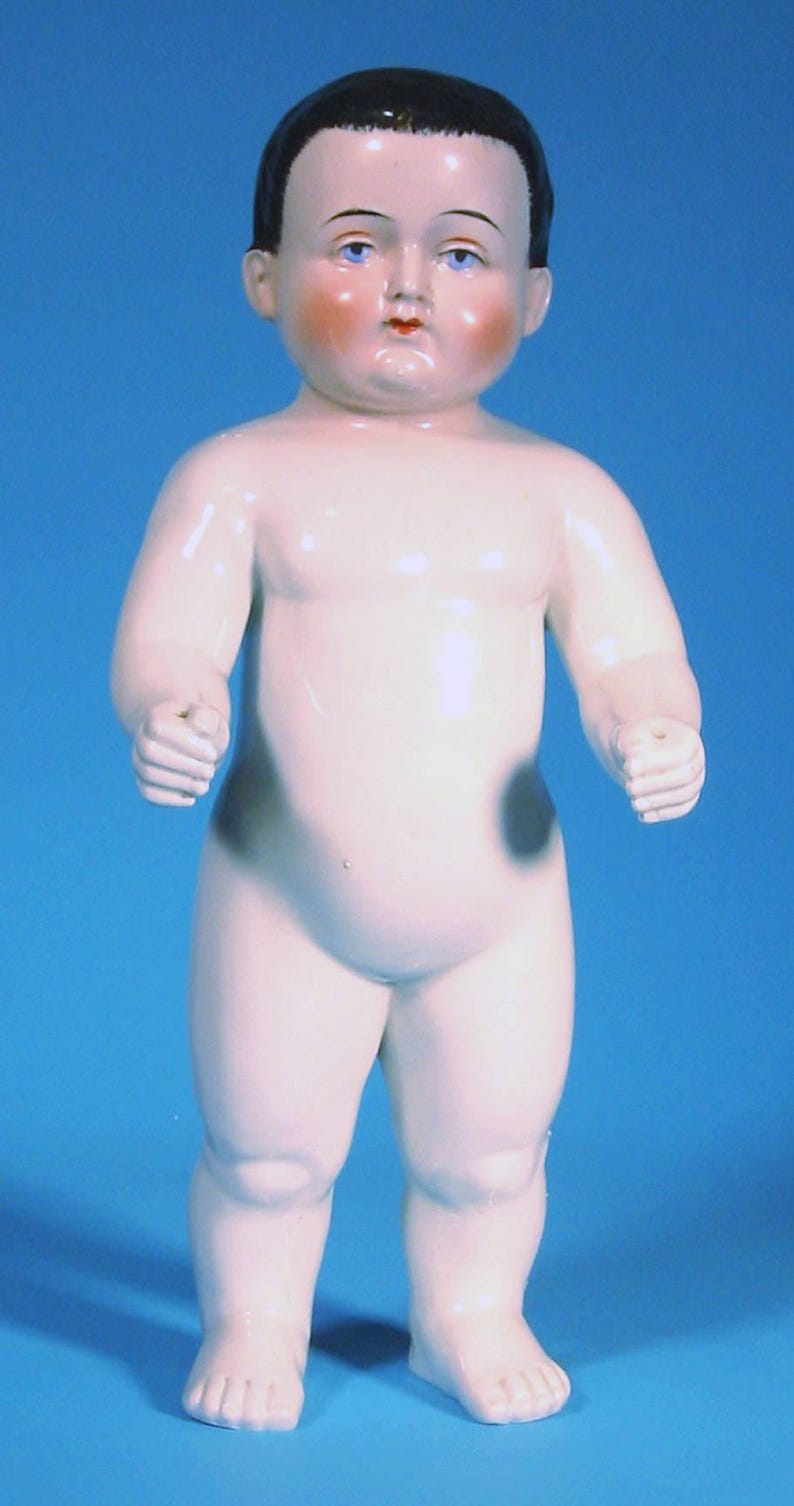 May include: A porcelain doll with a smooth, pale finish, dark painted hair, and blue eyes. The doll features rosy cheeks, a small red mouth, and two distinct dark gray circular markings on the hips. It stands in an upright position with arms slightly bent at the sides, showcasing a glossy, reflective surface texture throughout the entire figure.