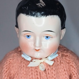 May include: A doll with a glossy porcelain head, painted black hair, and bright blue eyes. The doll features rosy cheeks, painted red lips, and a light blue collar. It wears a chunky, salmon-pink knitted sweater with a small white ribbon and button.