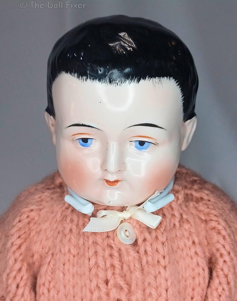 May include: A doll with a glossy porcelain head, painted black hair, and bright blue eyes. The doll features rosy cheeks, painted red lips, and a light blue collar. It wears a chunky, salmon-pink knitted sweater with a small white ribbon and button.