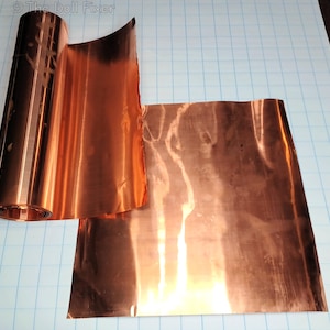May include: A roll of shiny copper foil next to a flat sheet of the same material. The copper has a reflective surface, and the roll is partially unrolled, revealing the foil's interior. The background is a light blue grid.