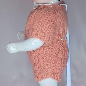 May include: A porcelain doll with black hair and a pale complexion, wearing a short-sleeved, peach-colored knitted romper. The doll is supported by a white metal stand with a round base. The text The Doll Fixer is visible in the top left corner.