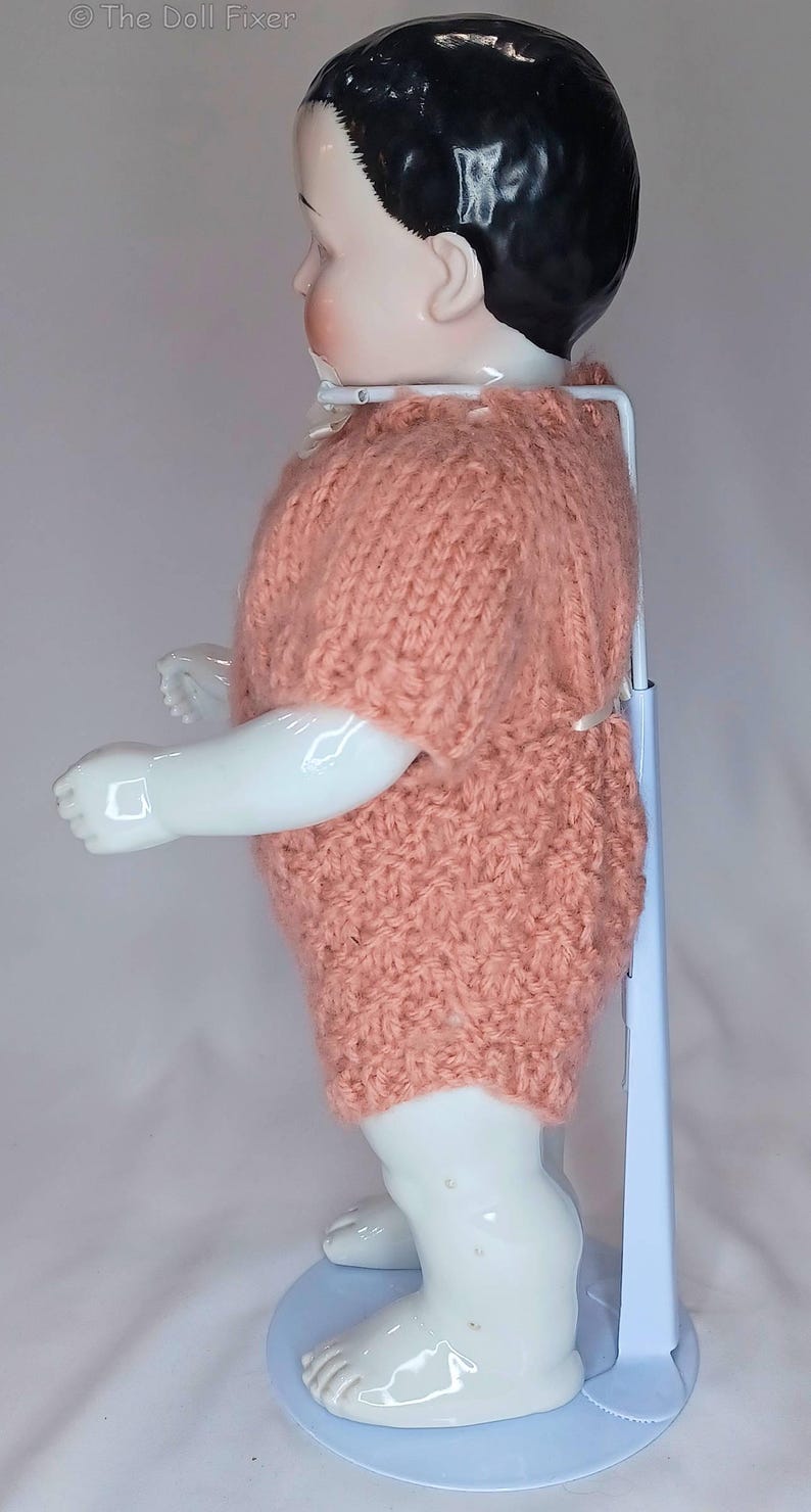 May include: A porcelain doll with black hair and a pale complexion, wearing a short-sleeved, peach-colored knitted romper. The doll is supported by a white metal stand with a round base. The text The Doll Fixer is visible in the top left corner.