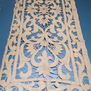 May include: Beige, openwork lace table runner with a floral and scroll design. The runner features a symmetrical pattern with detailed cut-out shapes and a scalloped edge. The fabric appears to be linen or a similar natural fiber.
