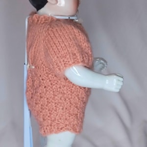 May include: A porcelain doll with black hair and a pale complexion, wearing a short-sleeved, textured peach-colored knitted romper. The doll is supported by a white metal stand and is viewed from the side, showing its smooth limbs and torso.