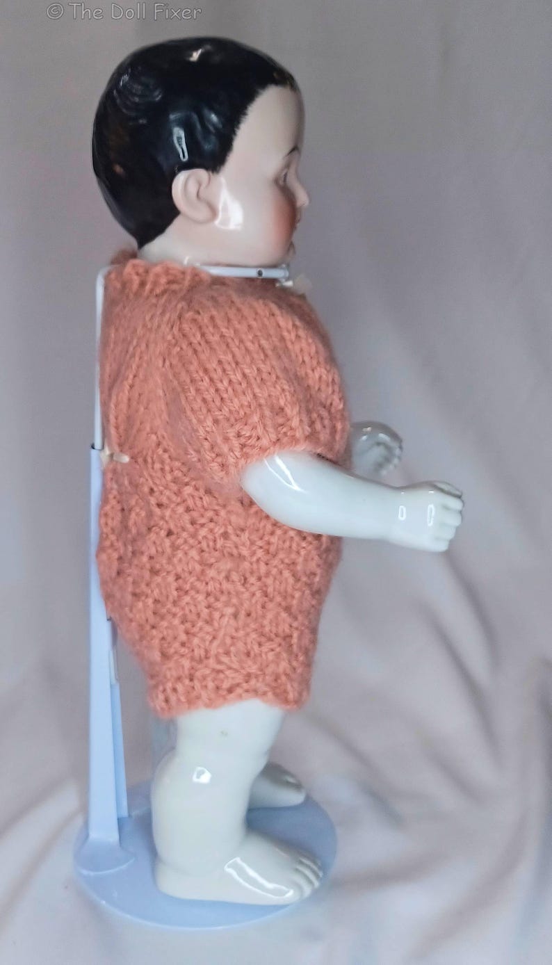 May include: A porcelain doll with black hair and a pale complexion, wearing a short-sleeved, textured peach-colored knitted romper. The doll is supported by a white metal stand and is viewed from the side, showing its smooth limbs and torso.