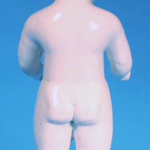 May include: A small, standing porcelain figurine of a baby with a smooth, white, glossy finish and molded dark brown hair. The figure is depicted from the back, showing a rounded body, short limbs, and a simple, unadorned, neutral appearance.