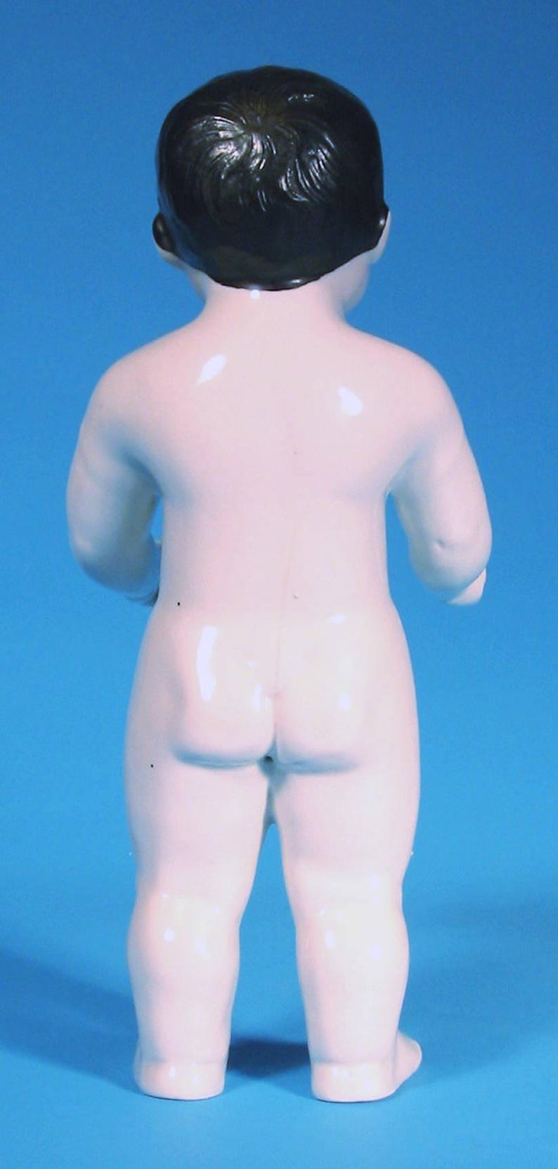 May include: A small, standing porcelain figurine of a baby with a smooth, white, glossy finish and molded dark brown hair. The figure is depicted from the back, showing a rounded body, short limbs, and a simple, unadorned, neutral appearance.