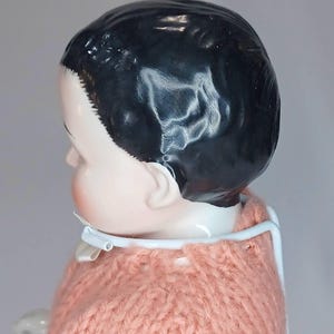 May include: A porcelain doll with a glossy black molded hairstyle and a pale complexion, wearing a peach-colored knitted sweater. The doll is supported by a white metal stand that encircles the neck. The text The Doll Fixer is visible.