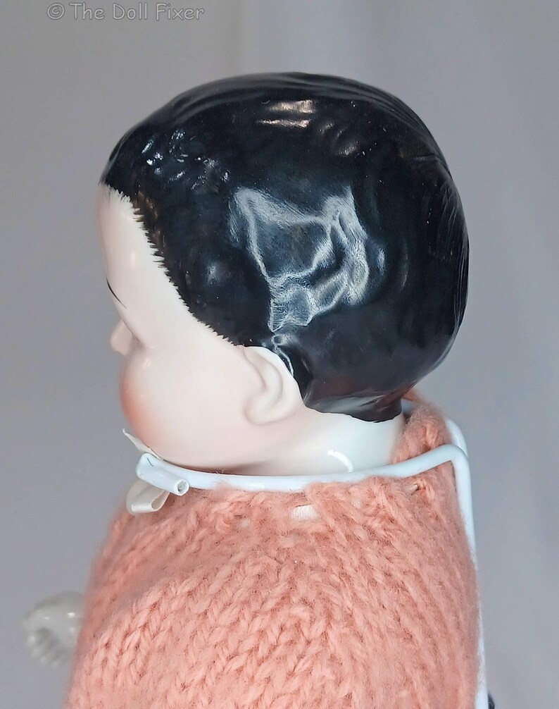 May include: A porcelain doll with a glossy black molded hairstyle and a pale complexion, wearing a peach-colored knitted sweater. The doll is supported by a white metal stand that encircles the neck. The text The Doll Fixer is visible.