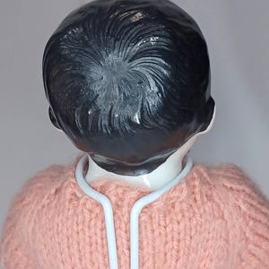 May include: A doll with a smooth, white ceramic head and molded black hair featuring a swirling texture. The doll is wearing a light pink, hand-knitted sweater and is supported by a white metal stand that curves around the neck area.