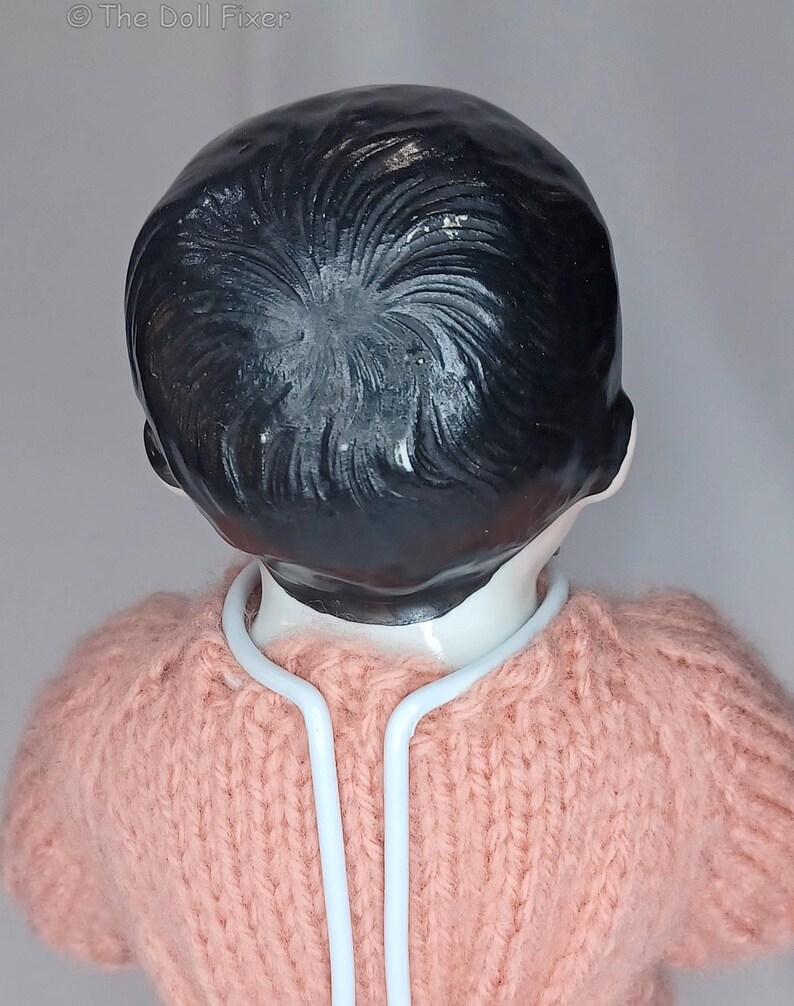 May include: A doll with a smooth, white ceramic head and molded black hair featuring a swirling texture. The doll is wearing a light pink, hand-knitted sweater and is supported by a white metal stand that curves around the neck area.