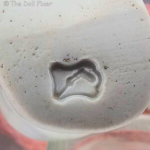 May include: A white ceramic surface featuring a small, irregular, recessed indentation in the center. The material has a speckled texture with tiny dark spots scattered across the off-white base. The text The Doll Fixer is visible in the top left corner.