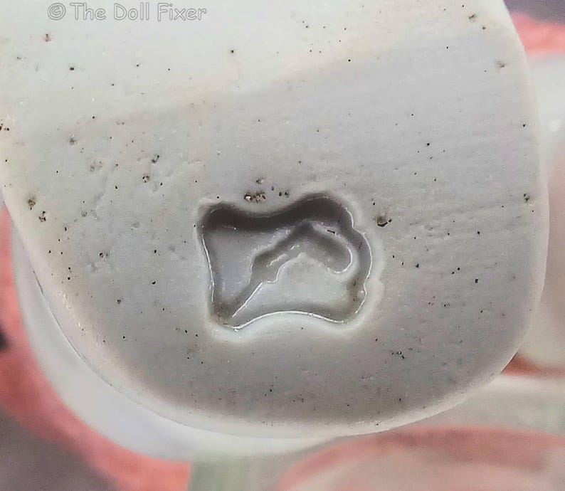 May include: A white ceramic surface featuring a small, irregular, recessed indentation in the center. The material has a speckled texture with tiny dark spots scattered across the off-white base. The text The Doll Fixer is visible in the top left corner.