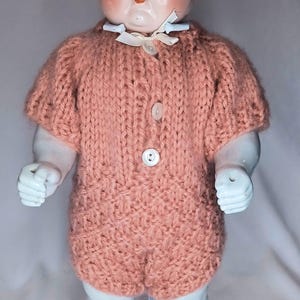 May include: A porcelain doll with painted black hair and blue eyes, wearing a short-sleeved, peach-colored knitted romper. The romper features a textured stitch pattern, a small white ribbon at the collar, and three white buttons down the front.